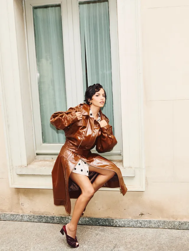 Isabela Merced sitting by a window in a brown coat and heels.