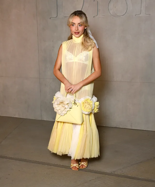 Sabrina Carpenter standing in a yellow gown and heeled sandals beside a wall.
