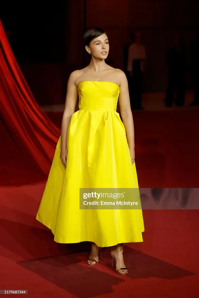 Naomi Scott posing in a yellow dress and gold heels.
