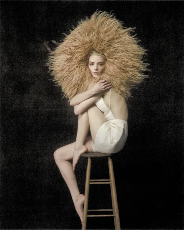 Anya Taylor-Joy seated barefoot on a wooden stool for a studio portrait.