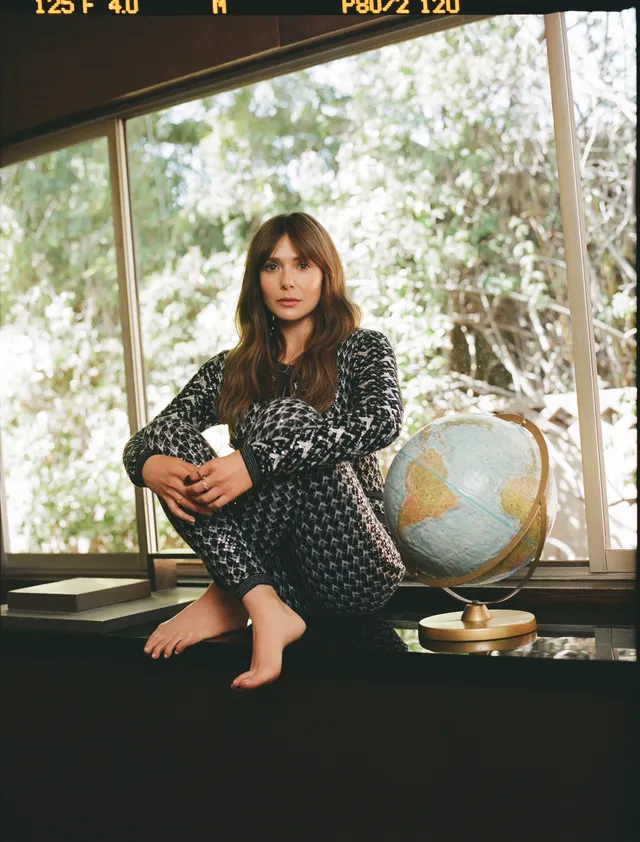 Elizabeth Olsen sitting barefoot beside a globe and window.