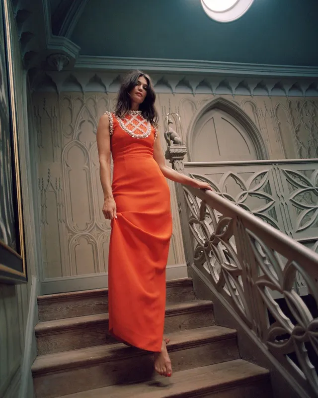 Gemma Arterton standing on a staircase in an orange gown.
