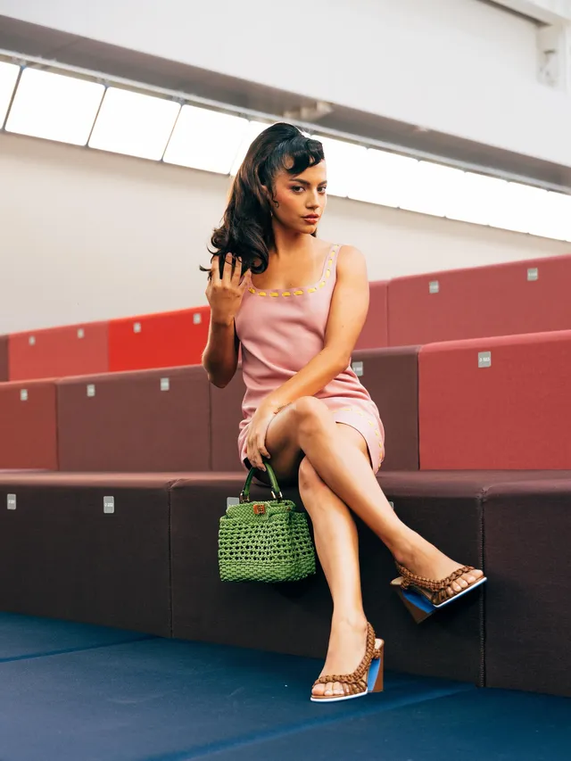 Isabela Merced sitting beside lockers in a pink dress and heeled sandals.