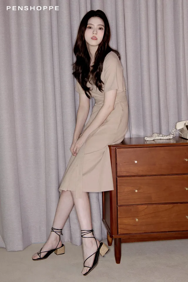 Han So-hee wearing a beige dress and black heels beside a dresser.