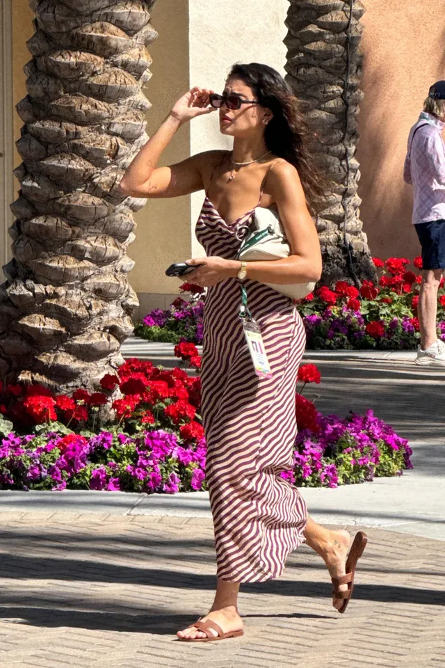 Eiza González walking outdoors in a striped dress and flat sandals.