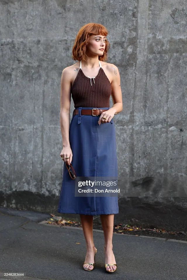 Ella Purnell posing beside a grey wall in a blue skirt.