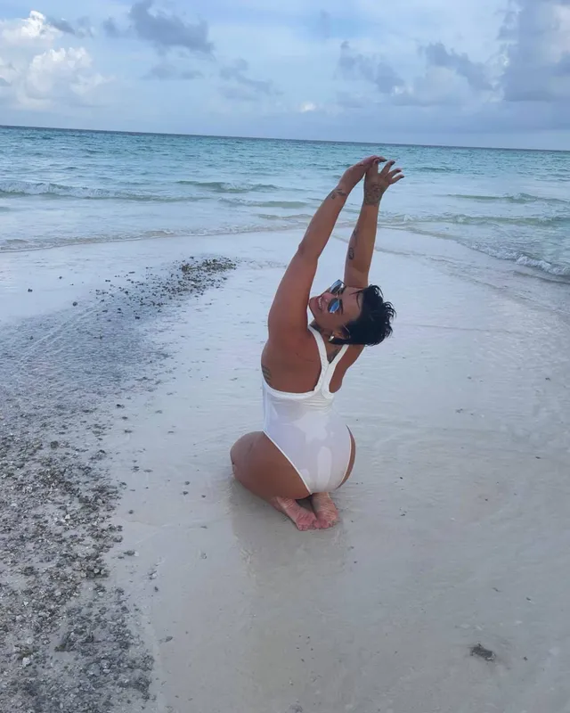 Demi Lovato kneeling barefoot on a sandy beach with arms raised.