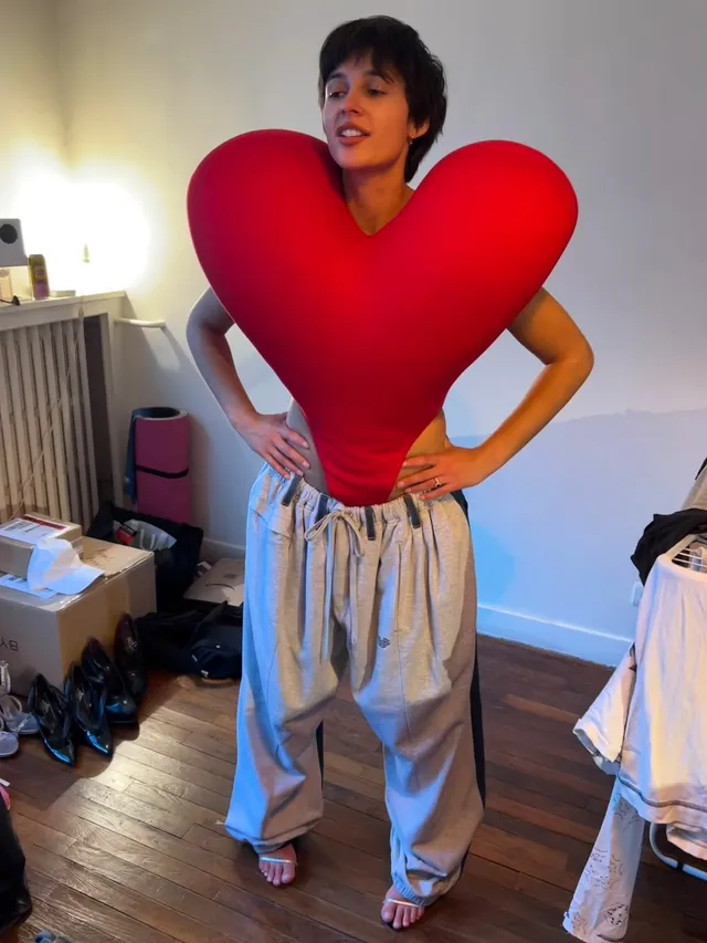 Naomi Scott posing indoors in pale trousers and heels.