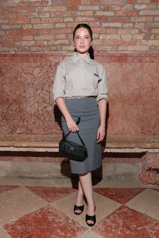 Cailee Spaeny posing beside a brick wall in a grey skirt and black heels.
