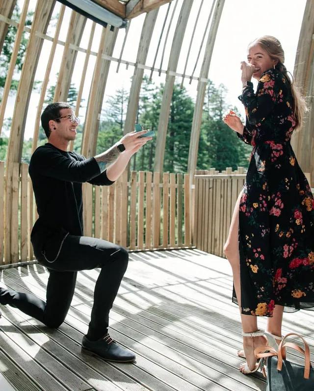 Debby Ryan standing in a floral dress and heels as a companion kneels nearby.