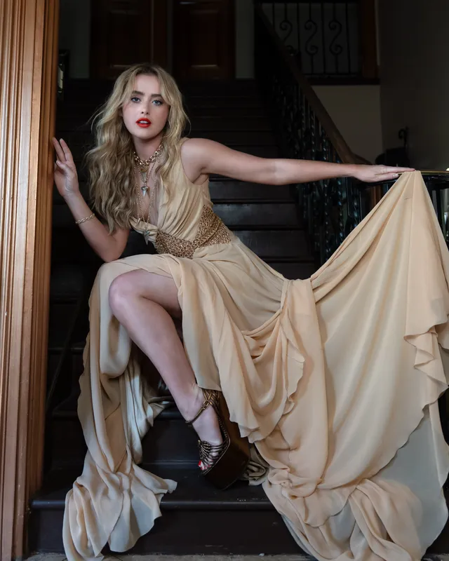 Kathryn Newton sitting on stairs in a gold gown and heeled sandals.
