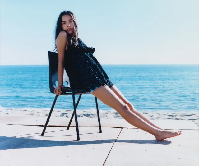 Olivia Rodrigo: seated barefoot on a chair beside the sea.