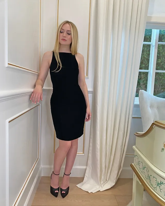 Dakota Fanning posing indoors in a black dress and heels.