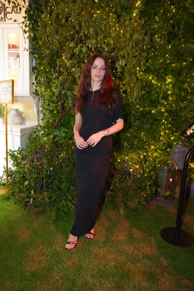 Joey King posing on grass in a black dress and heeled sandals.
