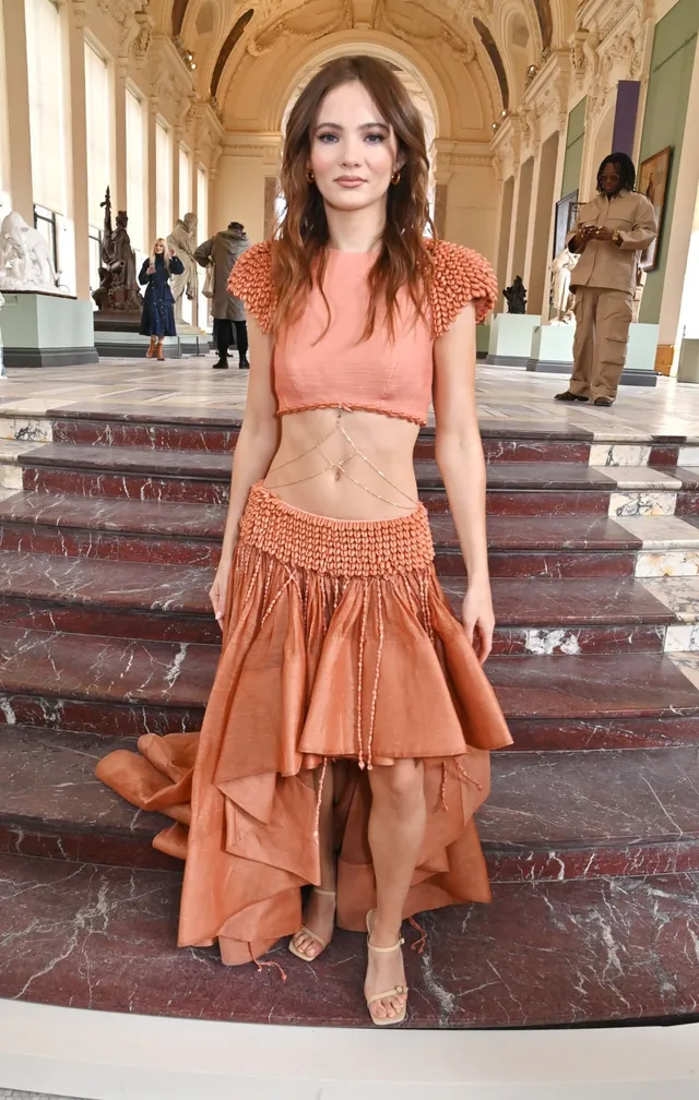 Freya Allan posing in an orange dress and pale heels on stairs.