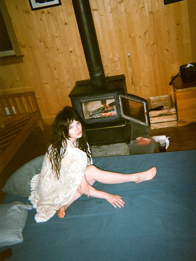 Sophie Thatcher sitting barefoot on a floor beside a wood-burning stove.