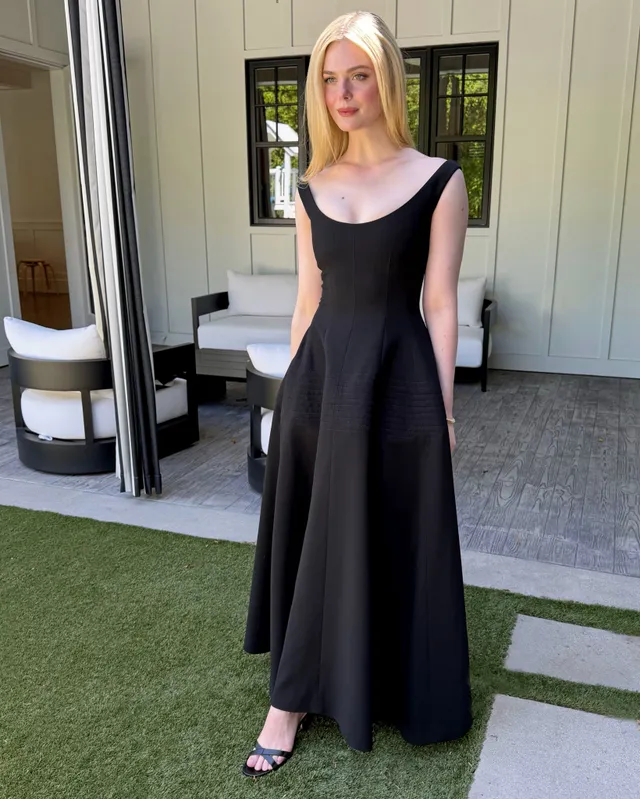 Elle Fanning posing in a long black dress and heeled sandals outside.
