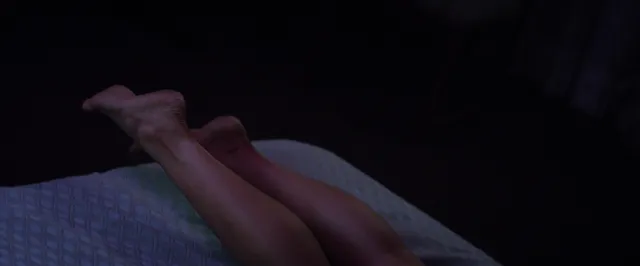 Megan Fox showing bare feet resting on bedding in a dim film scene.