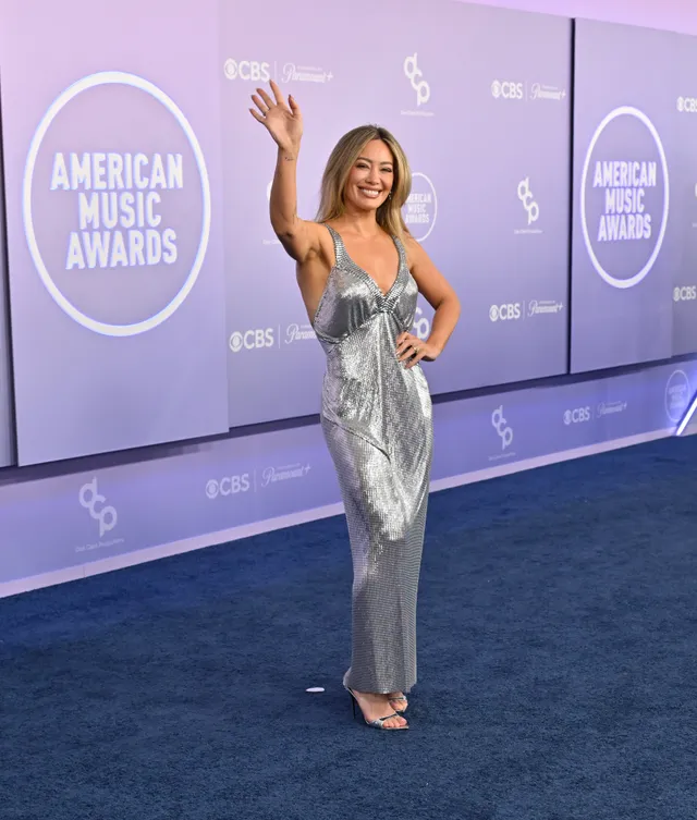 Hilary Duff waving in a silver dress and heels against an event backdrop.