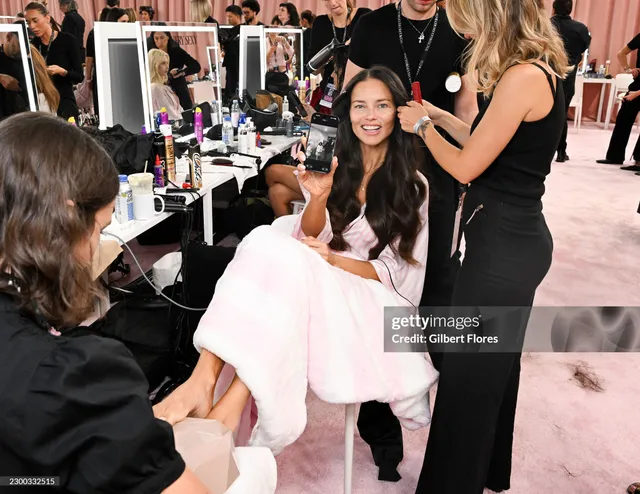 Adriana Lima seated barefoot backstage while a stylist works nearby.