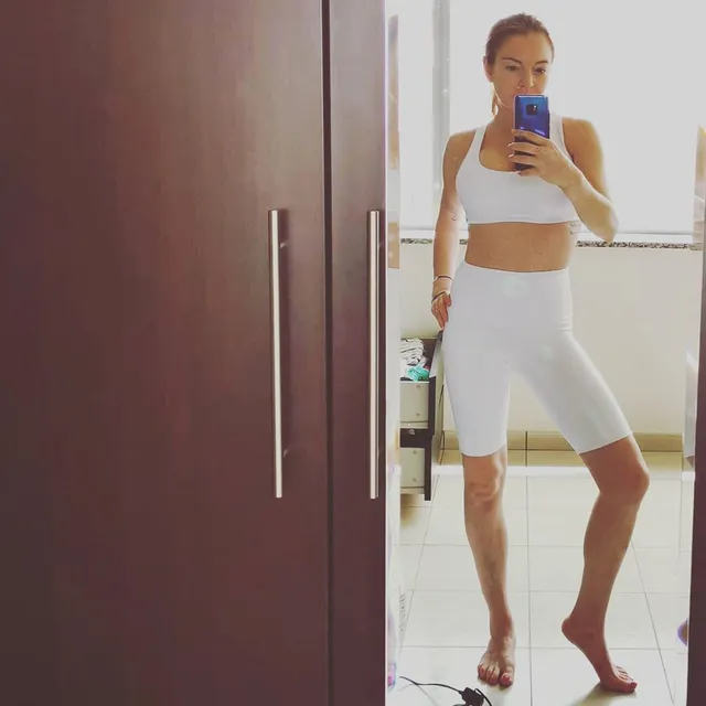 Lindsay Lohan standing barefoot in a white athletic outfit for a mirror photograph.
