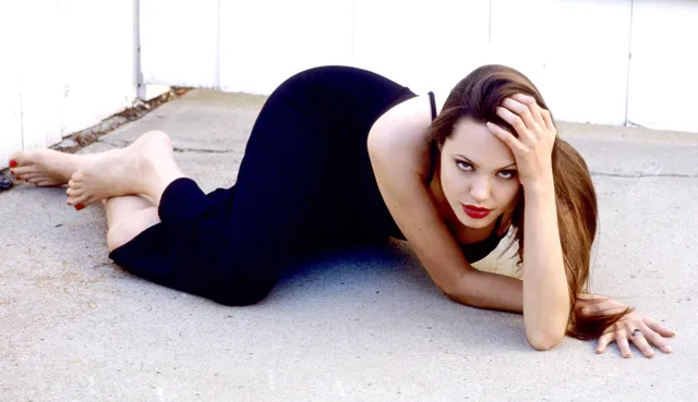 Angelina Jolie lying on a carpet in a black dress with her bare feet raised.