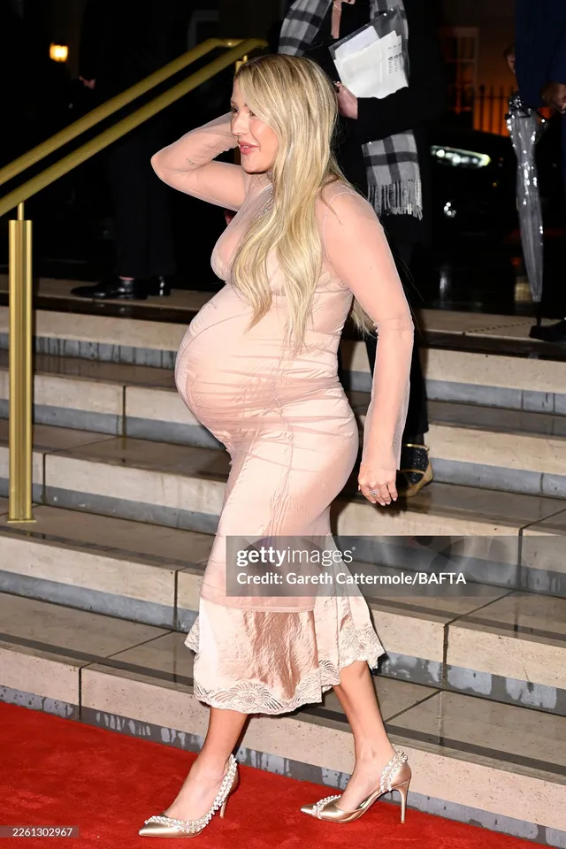 Ellie Goulding standing on a red carpet in a pale dress and white heels.