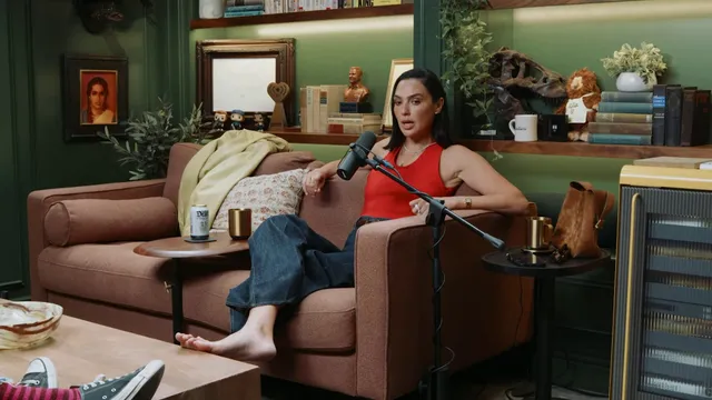 Gal Gadot sitting barefoot on a sofa during a conversation.