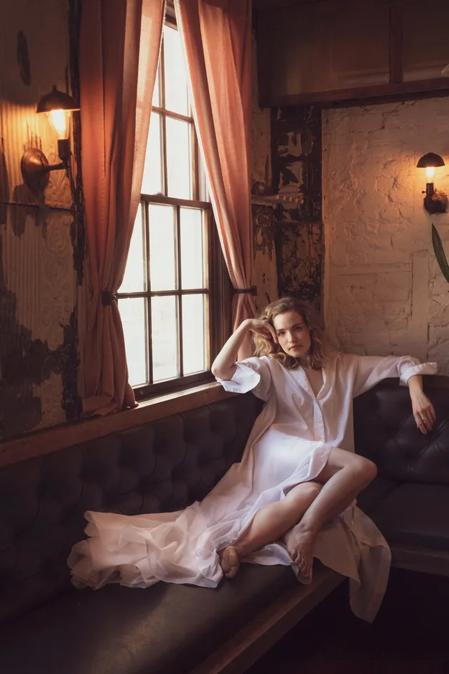 Willa Fitzgerald reclining barefoot by a window in a pale dress.