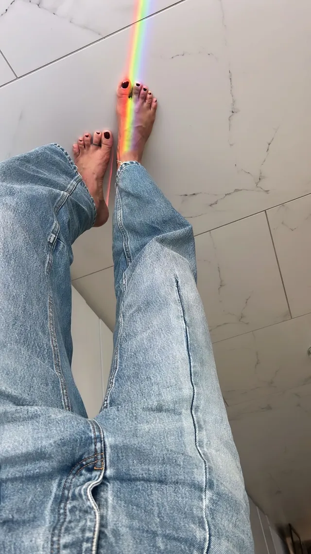 María Pedraza shown standing barefoot in jeans beside a stripe of rainbow light.