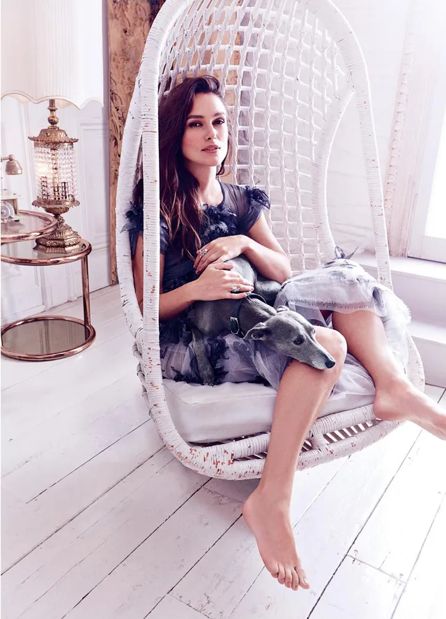 Keira Knightley relaxing in a white hanging chair in a softly lit room.