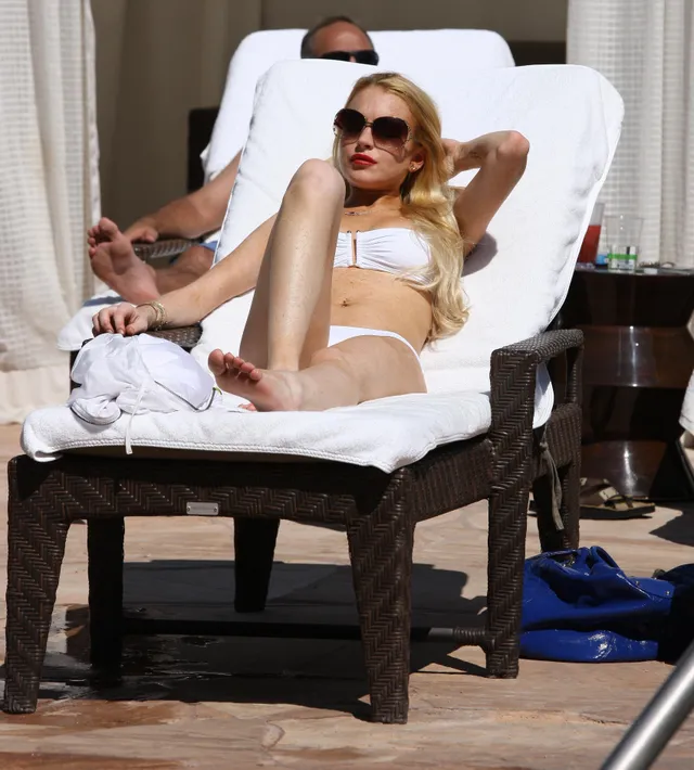 Lindsay Lohan relaxing barefoot on a white lounger in a white swimsuit.