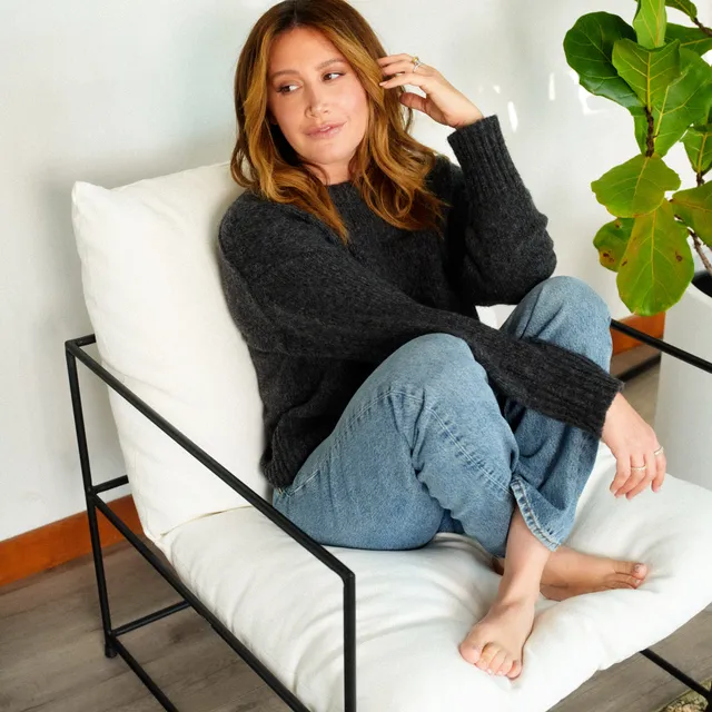 Ashley Tisdale sitting barefoot in a white chair wearing jeans and a dark sweater.