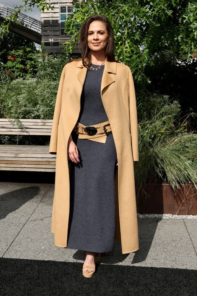 Hayley Atwell standing outdoors in a camel coat and sandals.