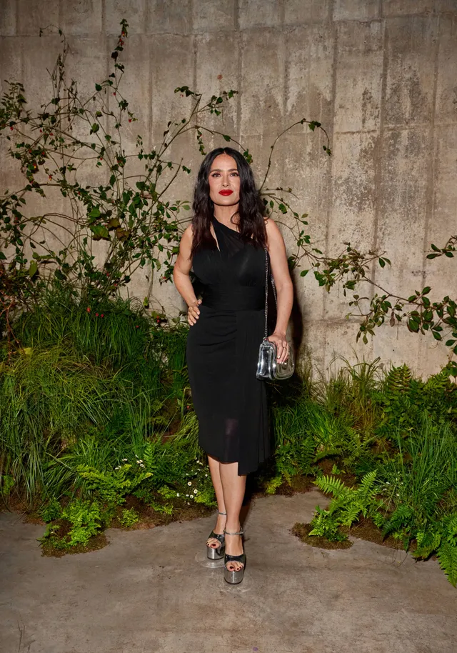 Salma Hayek posing outdoors in a black dress and heels.