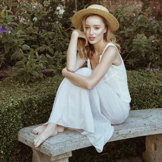 Phoebe Dynevor seated on a garden bench in a white dress and straw hat.