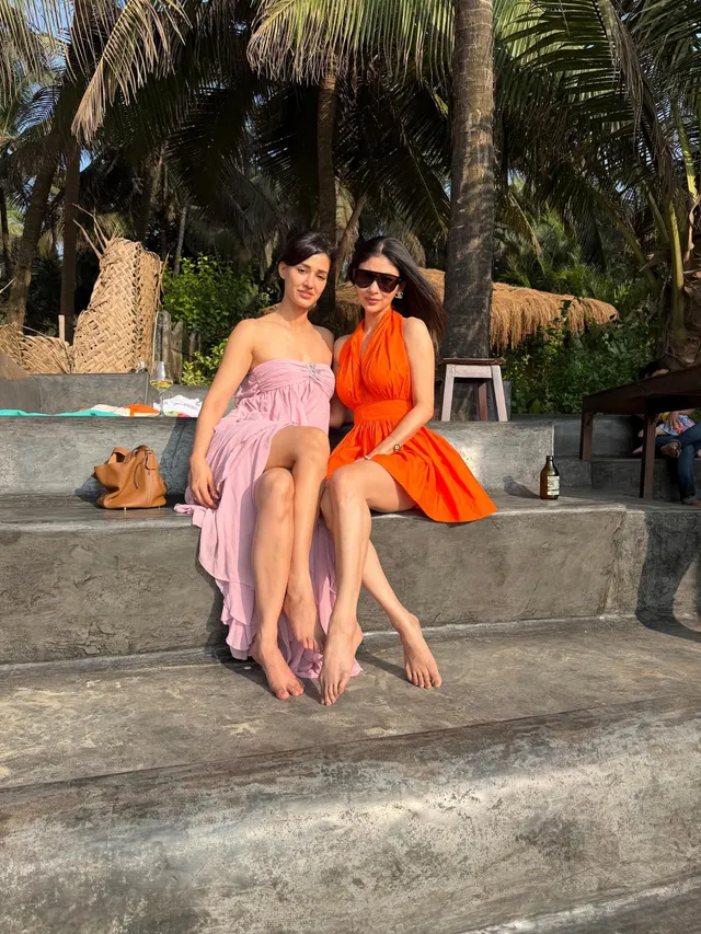 Disha Patani sitting barefoot beside a companion on stone steps beneath palm trees.