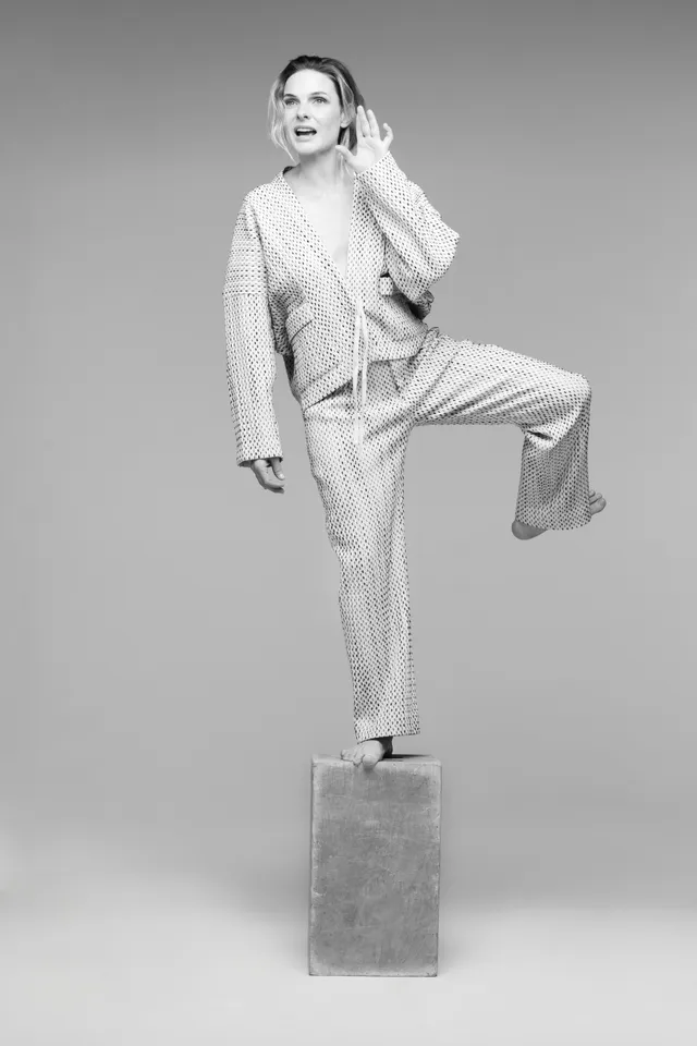 Rebecca Ferguson balancing on a pedestal in a tailored suit and heels in a monochrome portrait.