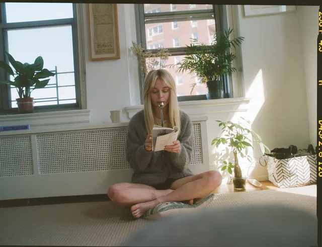 Emily Alyn Lind sitting cross-legged barefoot beside a window while reading.