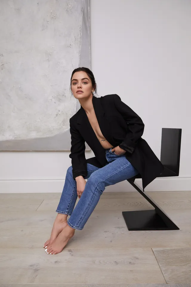 Lucy Hale sitting on a chair in a black jacket and jeans.