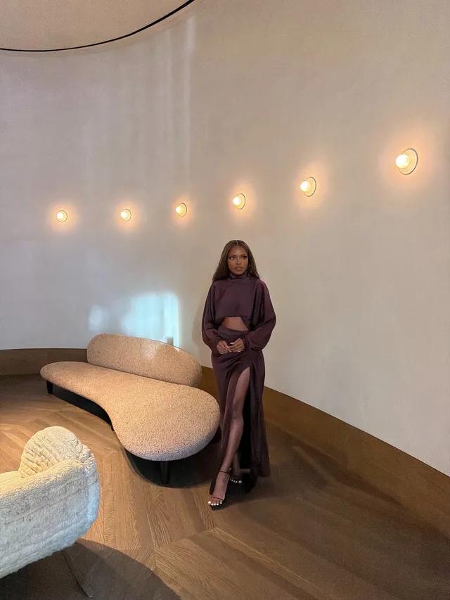 Ryan Destiny posing in a purple outfit and metallic heels beside a sofa.
