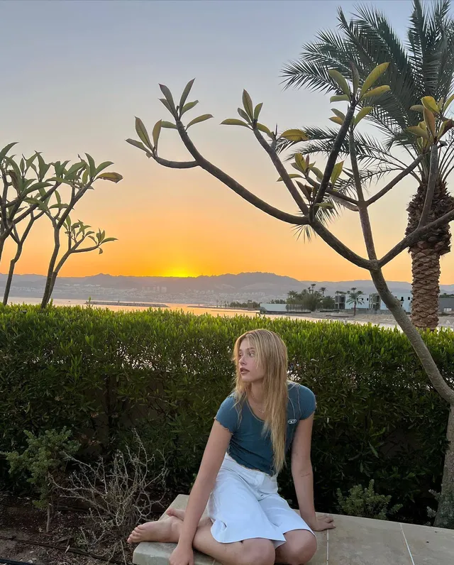 Nicole Wallace sitting barefoot outdoors in a blue top and light skirt at sunset.
