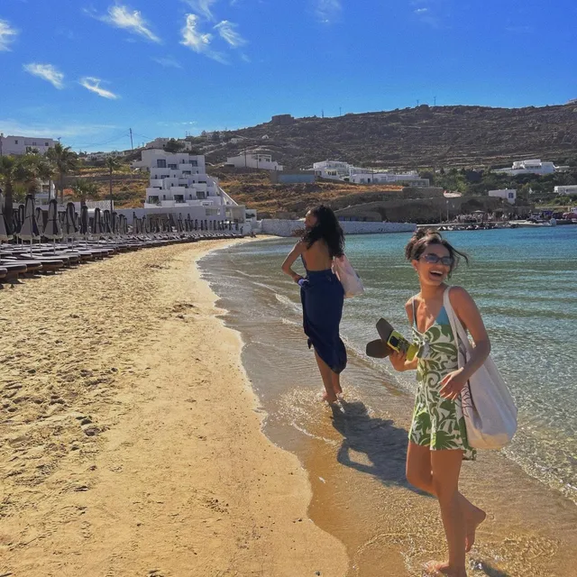 Rachel Zegler walking barefoot along the shore beside another woman.