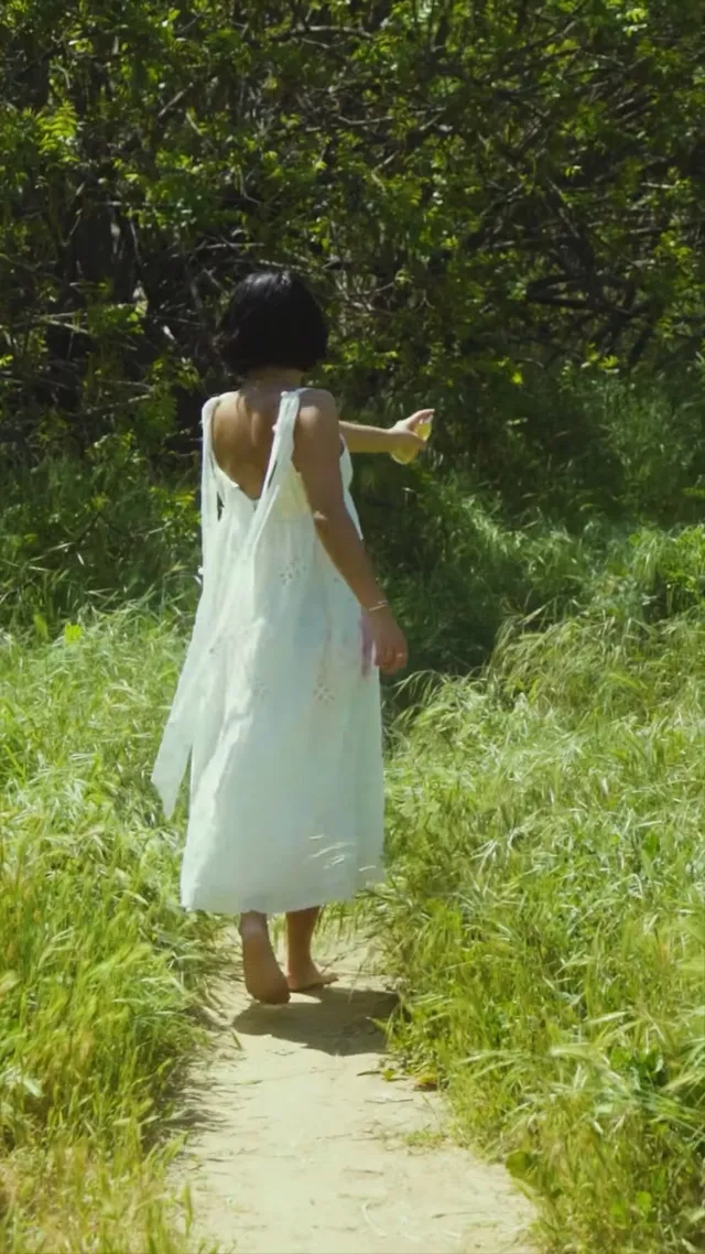 Isabela Merced standing barefoot on a garden path in a loose green dress.