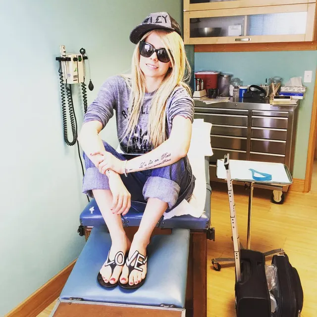 Avril Lavigne sitting in a room in cropped trousers and flip-flops.