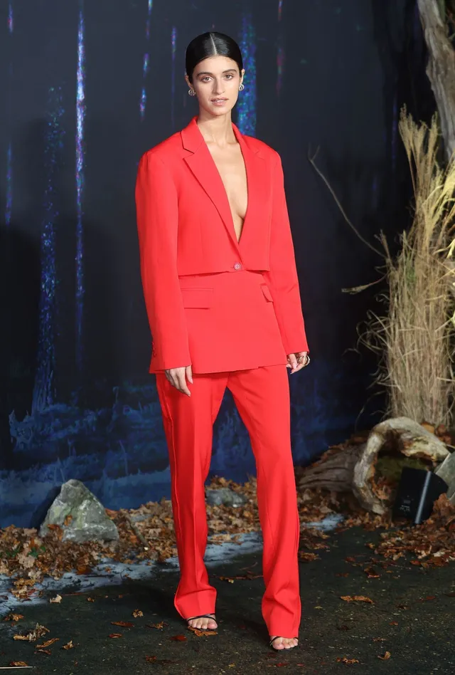Anya Chalotra standing in a red suit and dark heels at a premiere.