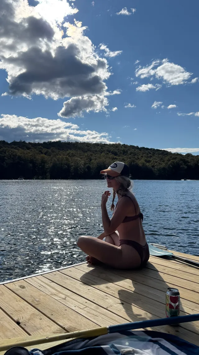 Brec Bassinger sitting barefoot on a wooden dock in a dark bikini.