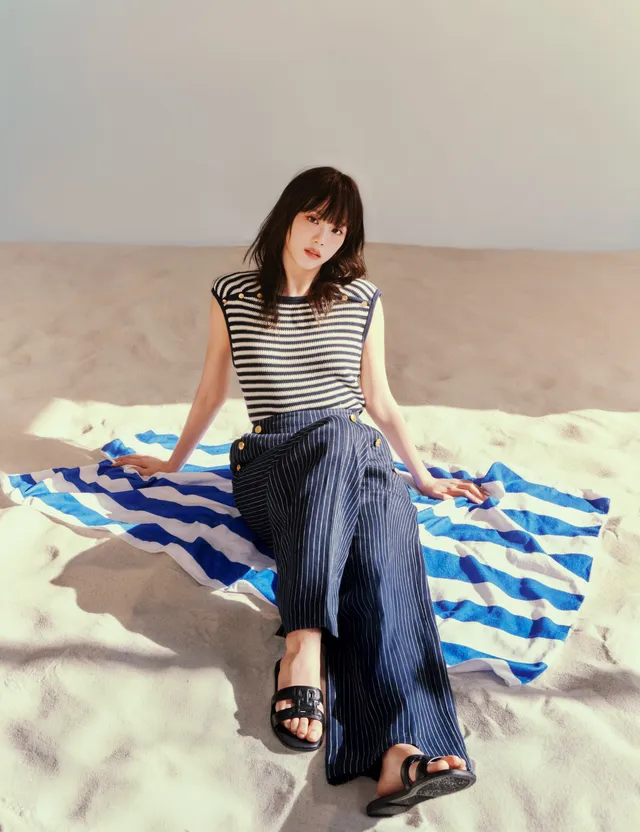 Jisoo sitting on a towel in black flat slides, striped top and dark trousers.