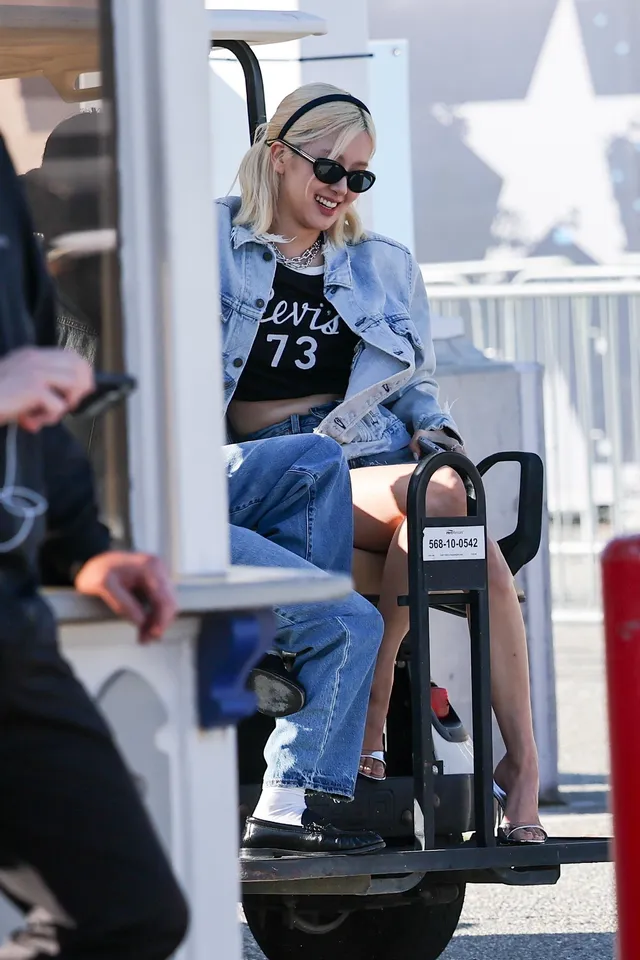 Rosé sitting on an airport cart in sunglasses, a denim jacket and jeans, with open-toe footwear visible.