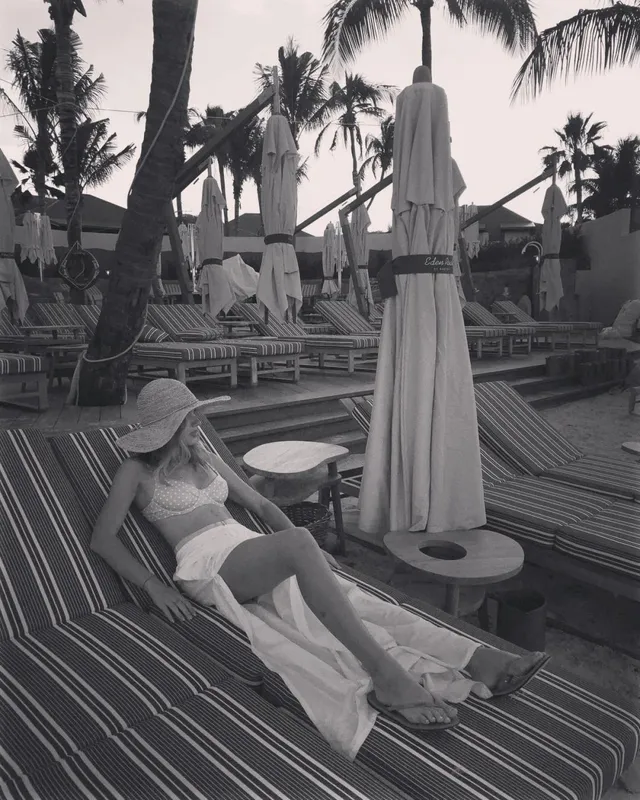 Halston Sage reclining barefoot on a pool lounger in a monochrome photograph.